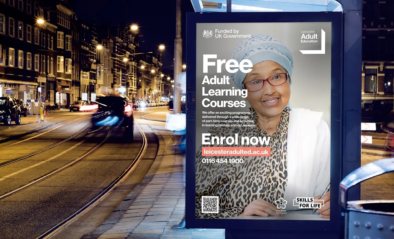 A bus stop advertisement at night, promoting free adult learning courses offered by Leicester Adult Education, funded by the UK Government. The ad features a woman wearing glasses and a headscarf, with text encouraging people to “Enrol now.”