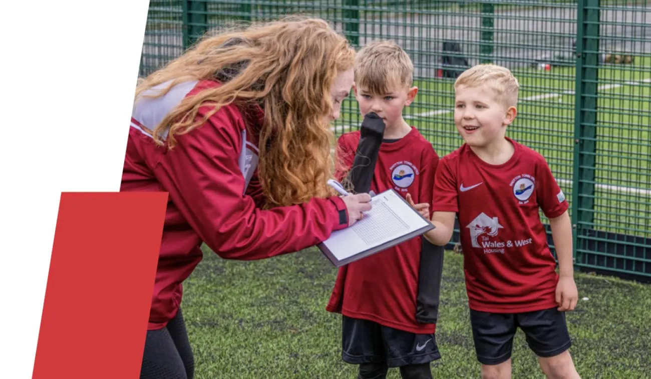 An image featuring a coach in a red jacket writing on a clipboard while interacting with two smiling young boys in football kits on a pitch.