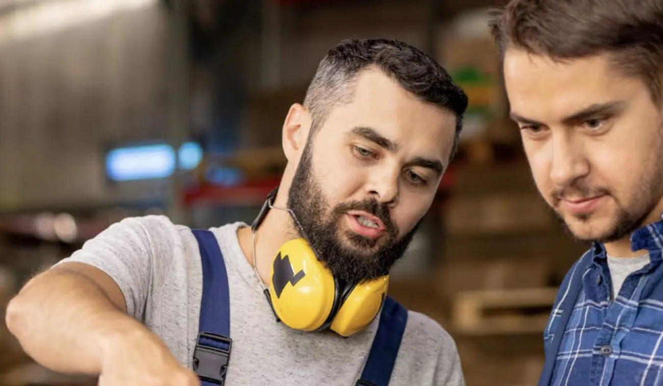 Two professionals collaborating in a workshop setting. The man on the left, wearing yellow protective headphones, gestures as he explains something to his colleague on the right, who listens attentively in a casual blue checkered shirt.