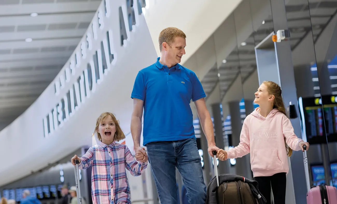 A father and his two daughters are seen smiling as they walk through an airport terminal, each pulling their own suitcase.