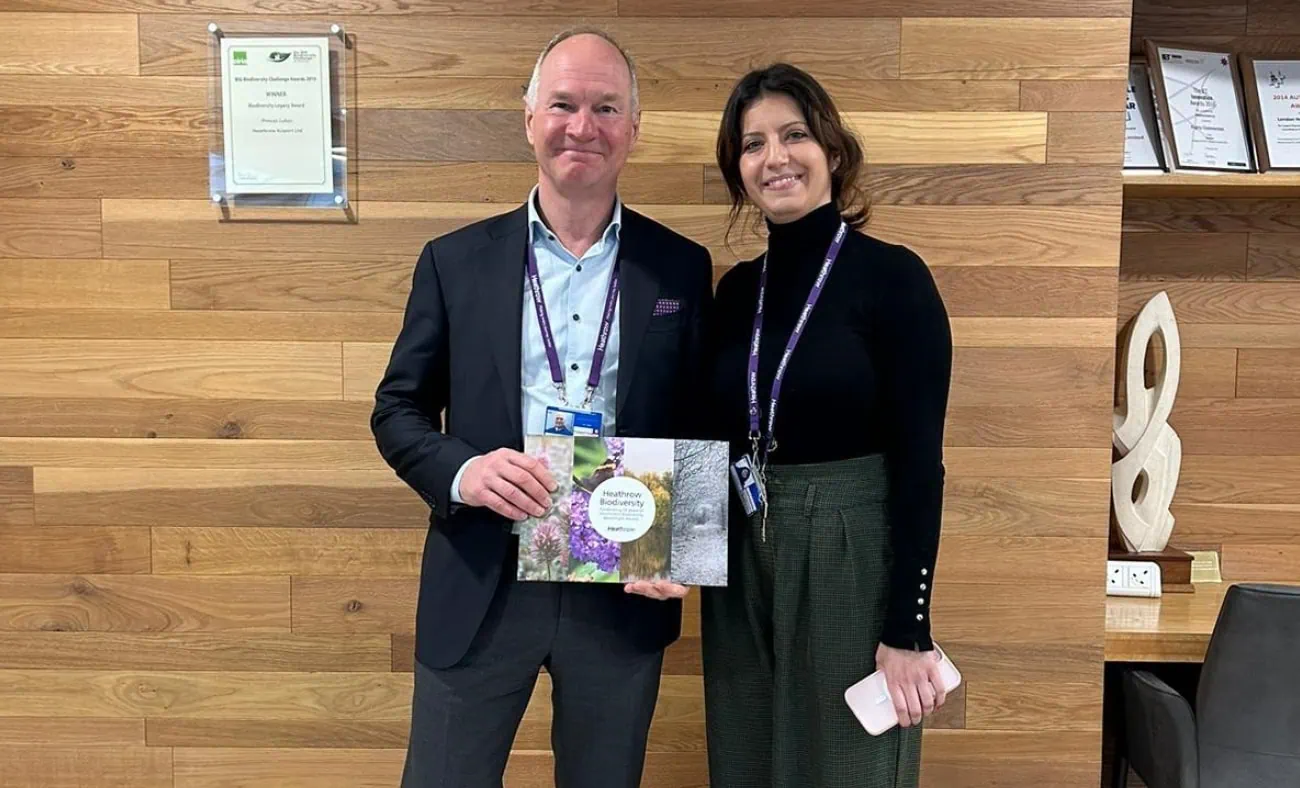 Two individuals stand indoors in front of a wooden-panelled wall. One is Heathrow CEO, Thomas Woldbye, who holds the Heathrow Biodiversity book.