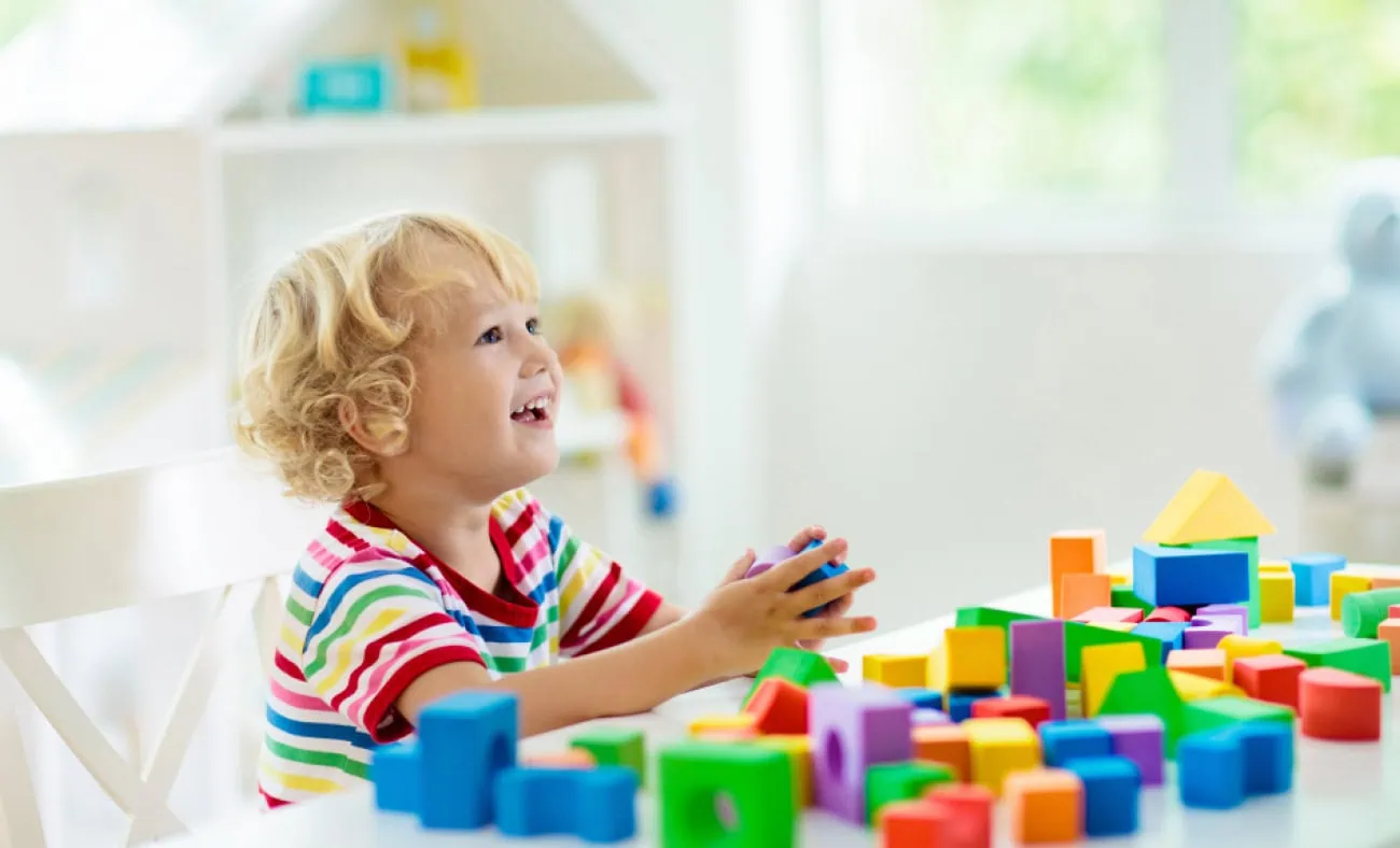 A young child sitting at a table, smiling and playing with colorful building blocks in a bright and airy room.