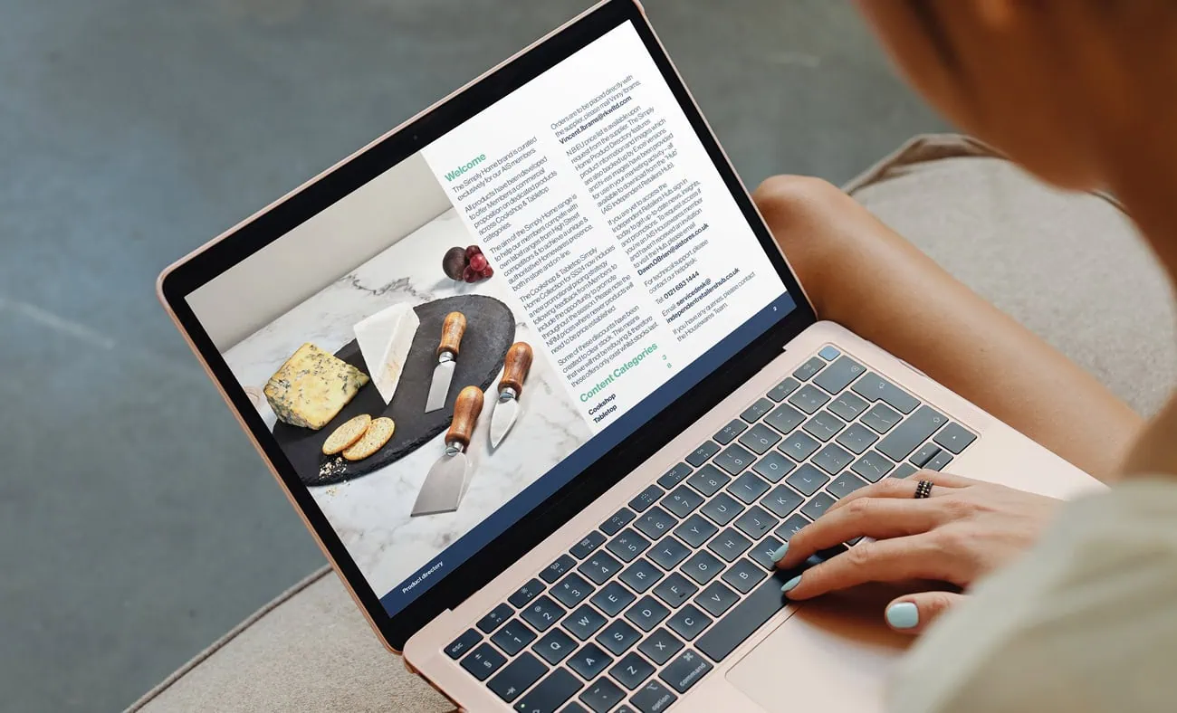 A person using a laptop to view a digital catalog page featuring a stylish cheese board display with various cheeses, knives, and accompanying text details.