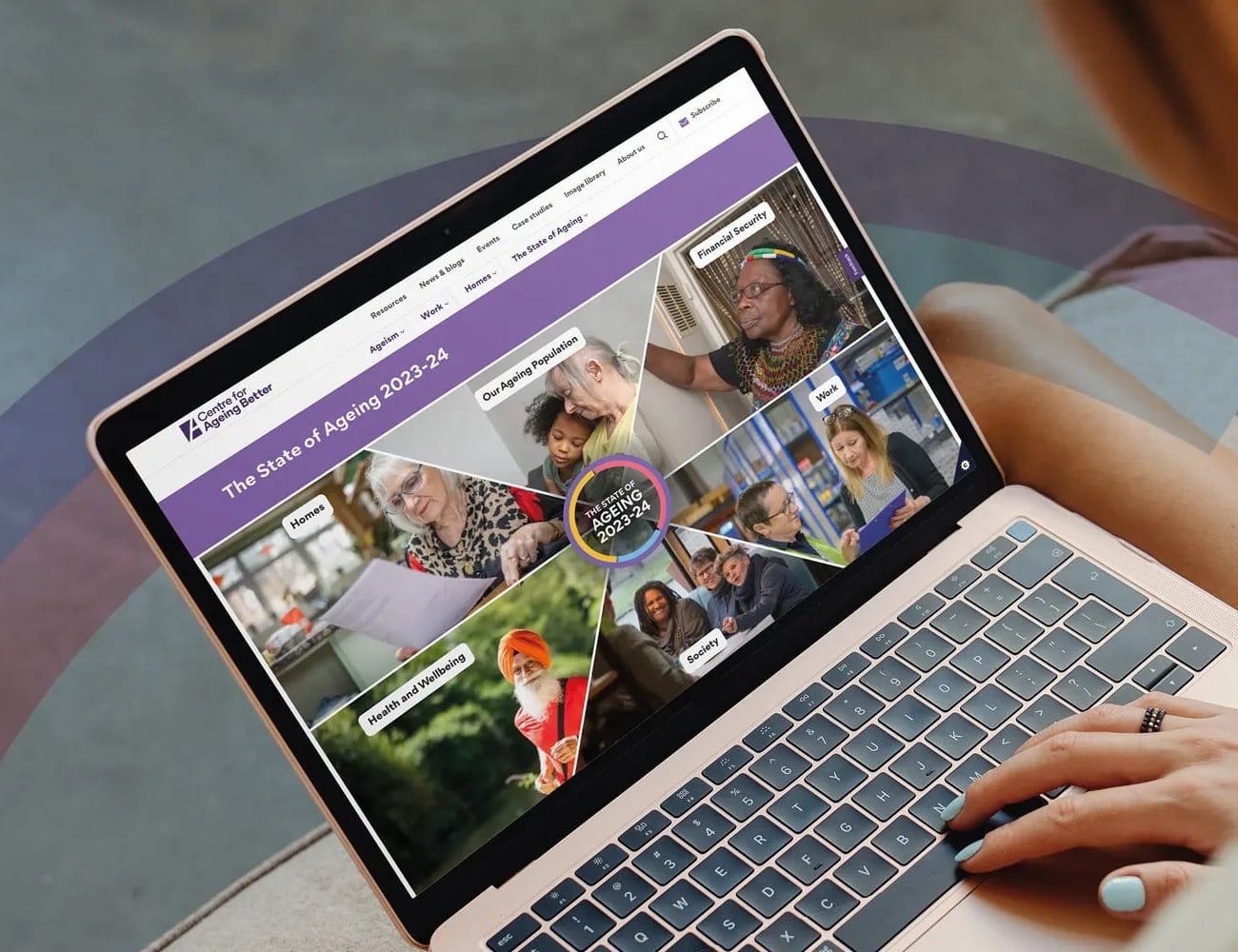 A person using a laptop displaying The State of Ageing 2023-24 website for Centre for Ageing Better. The screen features a segmented circular design with diverse images of older adults engaging in different activities, such as reading, working, and socialising.